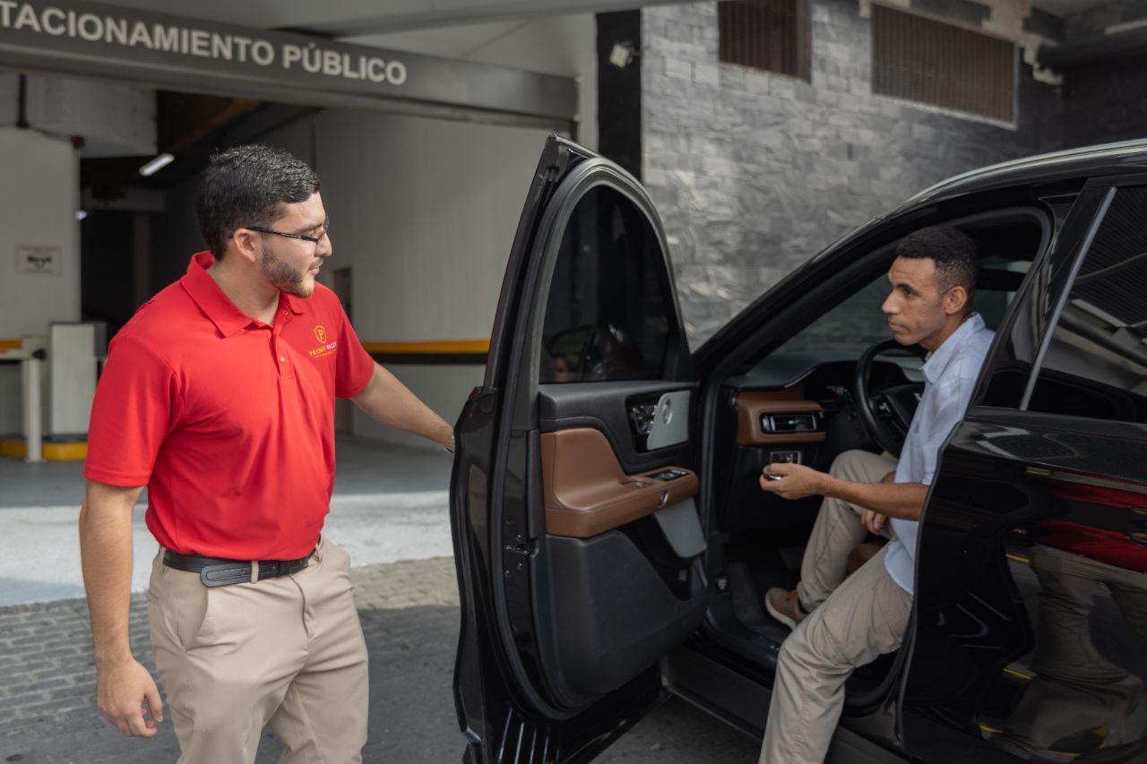 Valet en uniforme rojo Prime Valet recibe al cliente al abrir la puerta del vehículo en el estacionamiento público.