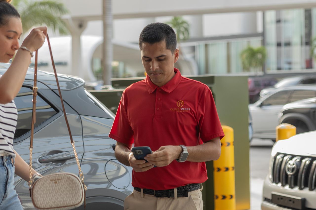 Valet Prime Valet en uniforme rojo revisando el registro en su teléfono junto a un vehículo gris en el acceso.